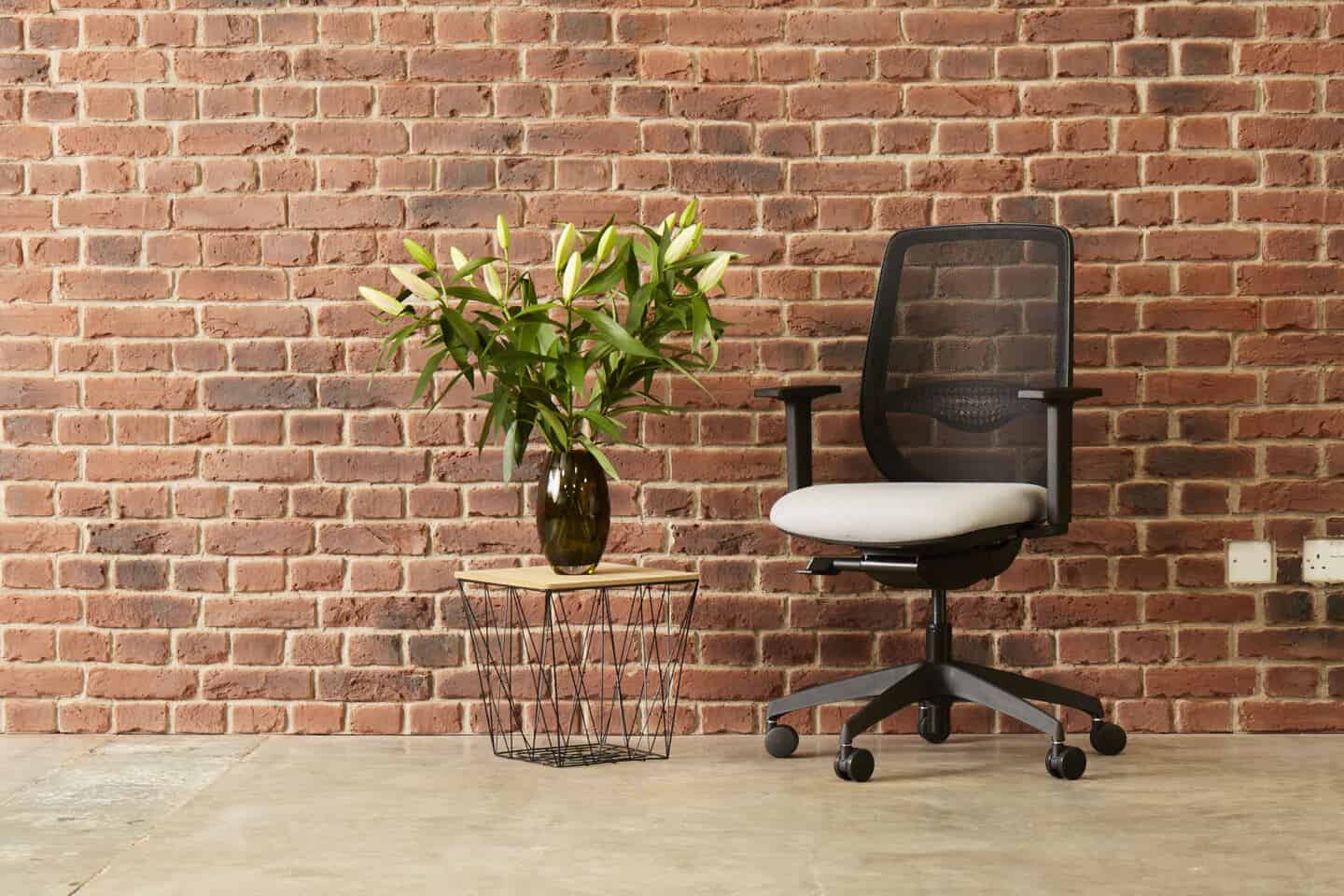 An ergonomic office chair infront of an exposed brick wall and next to a coffee table with a vase of flowers on it