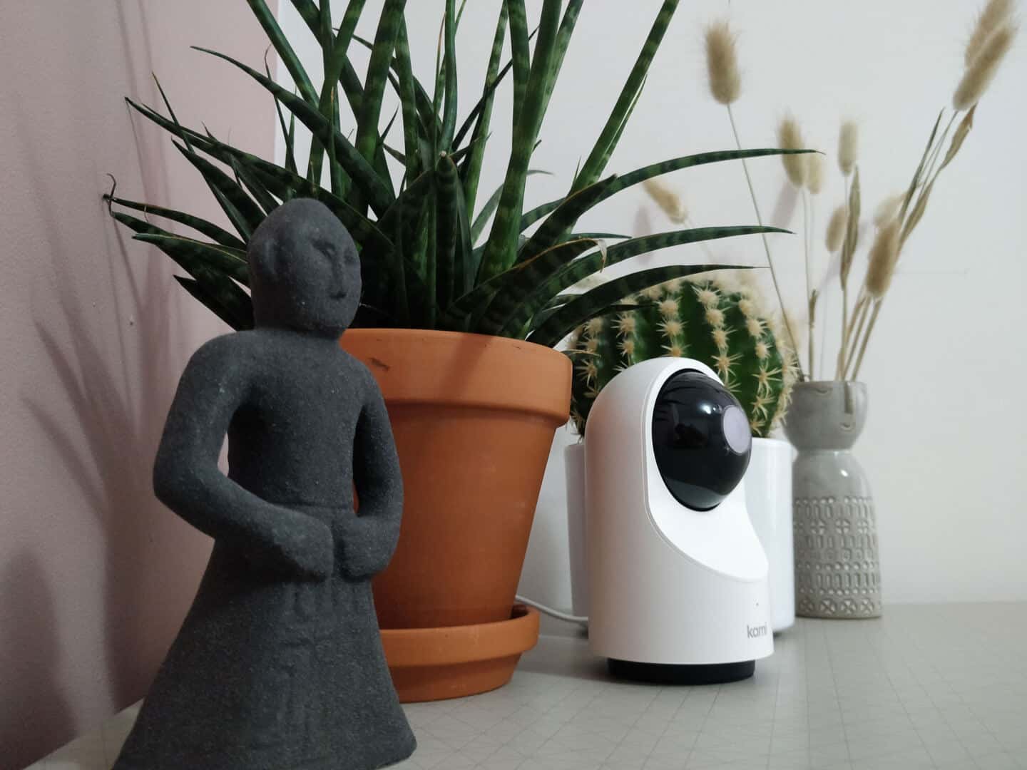 Kami indoor home security camera on a table between two plants and vase and a statue.