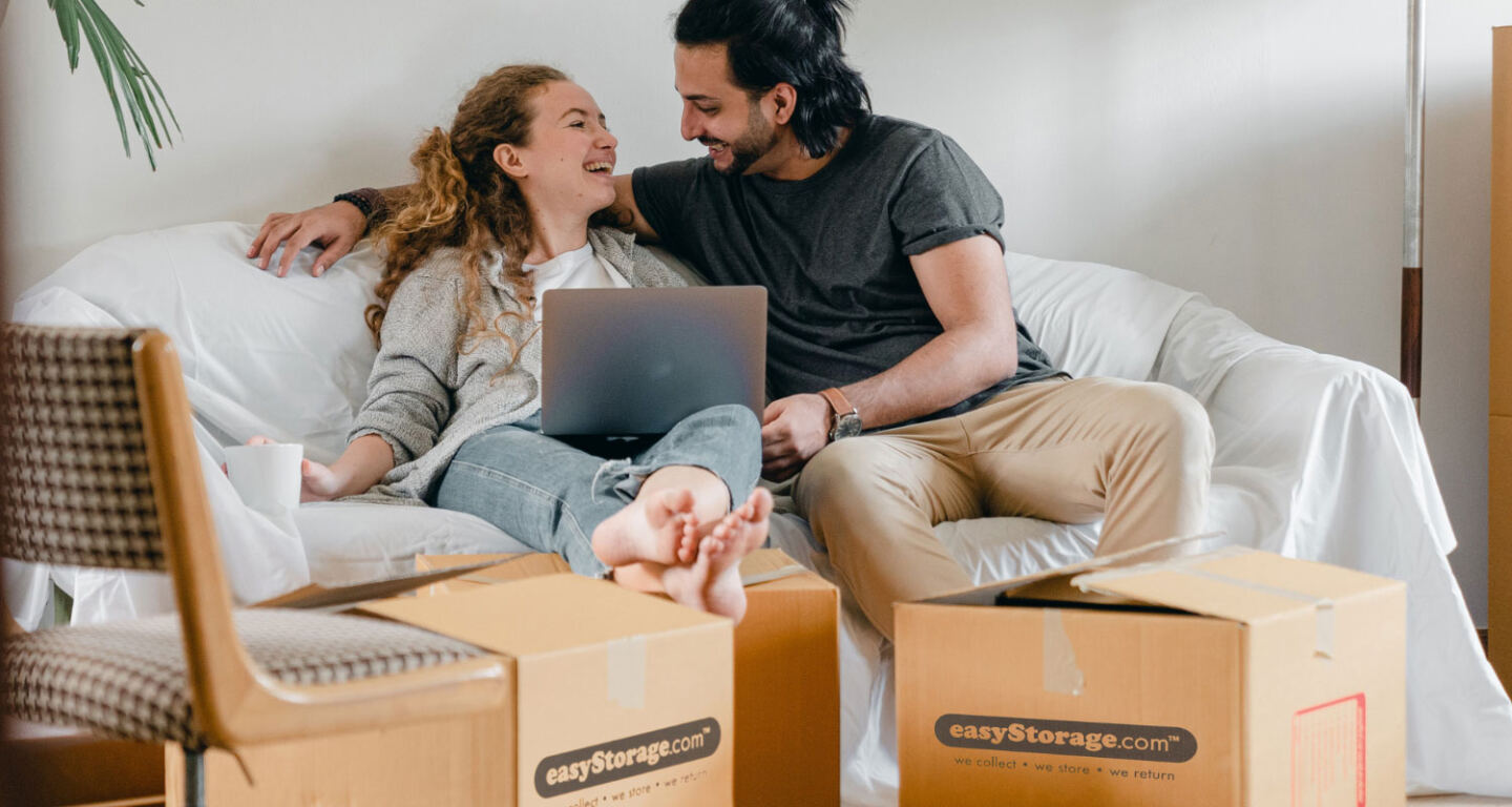 A young couple sit on a sofa with a laptop surrounded by cardboard boxes