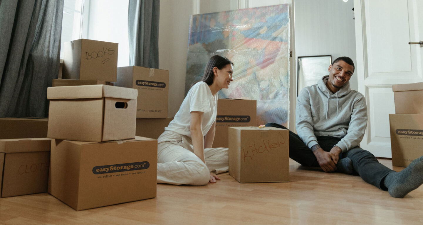 A young couple sat on the floor surrounded by cardboard boxes that are packed up with their belongings