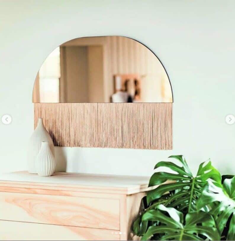 A semi circular mirror with fringing below hangs on a sage green wall with a dresser beneath and a monstera plant beside