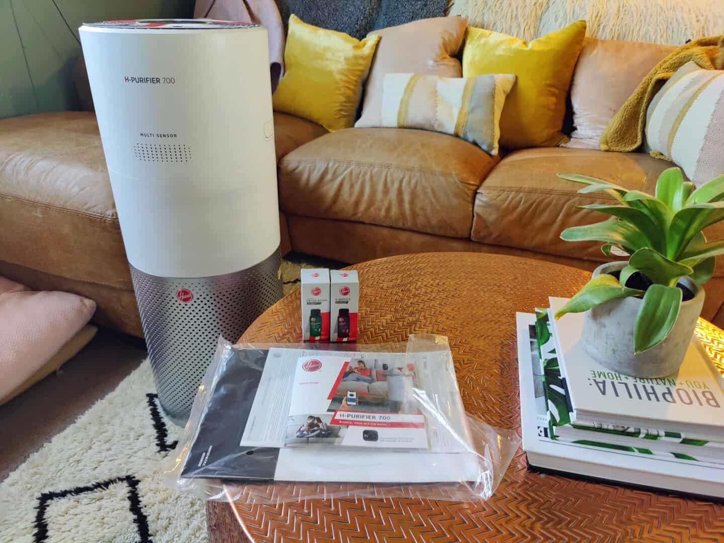 The Hoover Air Purifier 700 stands in a living room in front of a sofa and beside a coffee table. On the coffee table is the user manual, a pile of books and a plant.