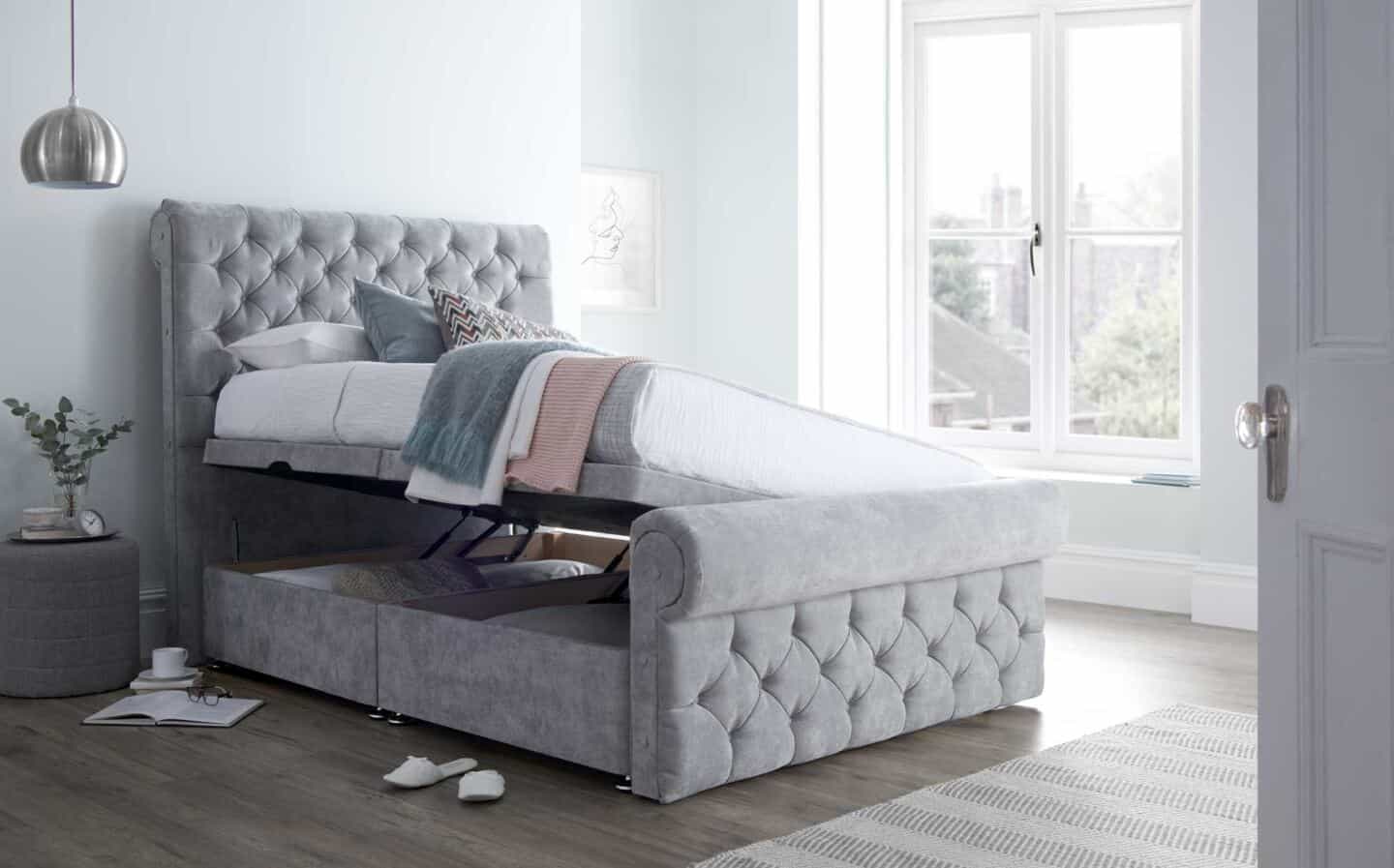 An open grey upholstered ottoman bed   in a bright an airy bedroom with large windows