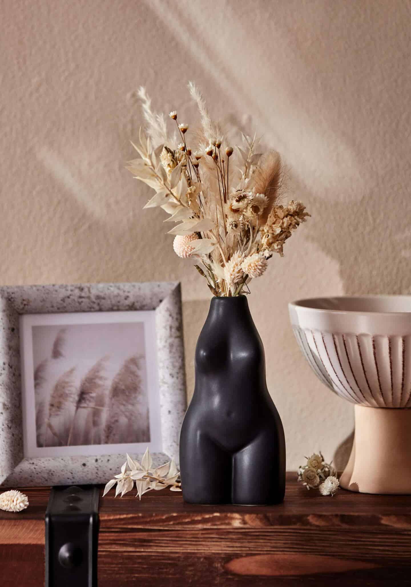 A small black vase in the shape of a female body sits besides a small photo frame and a small textured white bowl on a wooden shelf. 