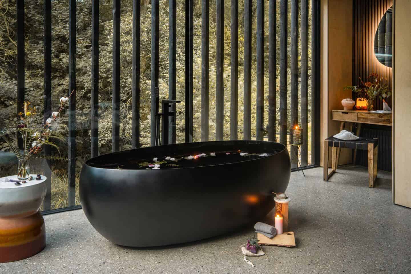 A large black freestanding bathtub in front of a large floor to ceiling window that overlooks the forest surrounding wildwood spa in north devon