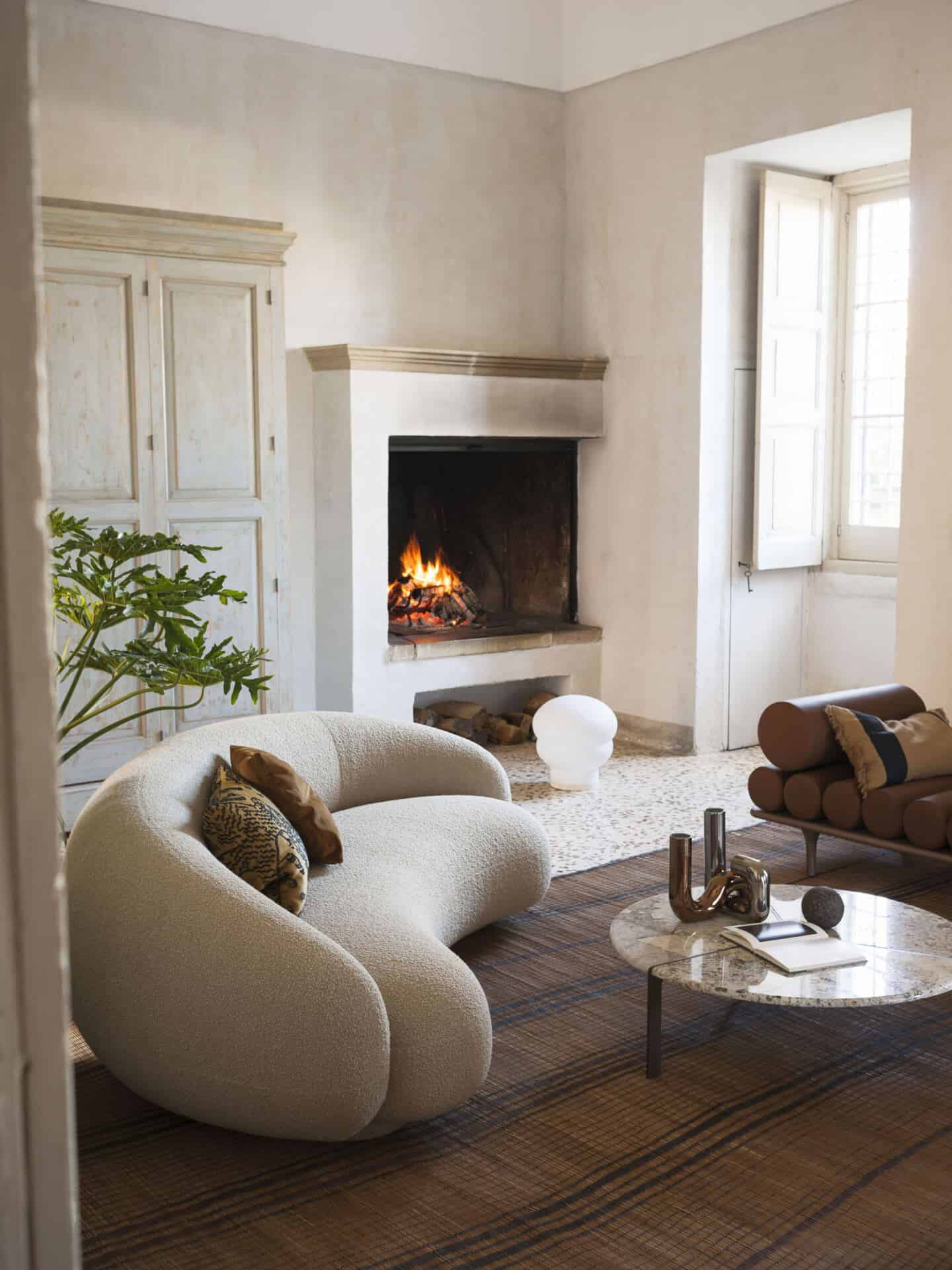 A curved bouclé sofa with an open fire behind and a marble coffee table in front