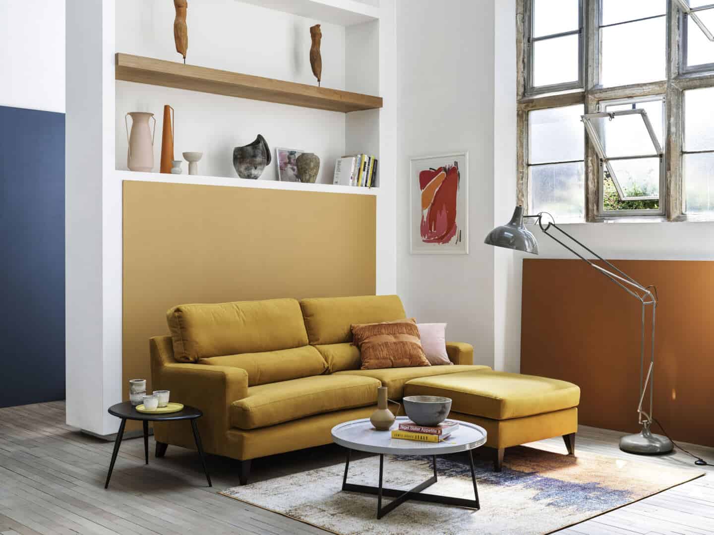 A yellow mid-century modern sofa with built-in shelving behind and industrial windows to the side.