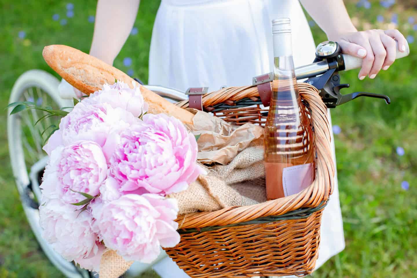 A bunch of pale pink peony flowers lies beside a baguette and a bottle of wine in the basket of a bicycle being ridden by a young white woman.