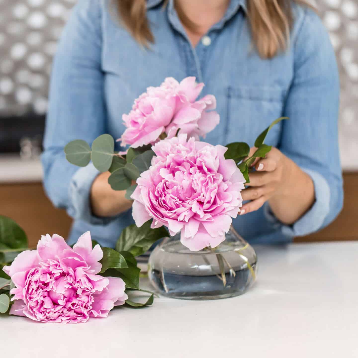 A woman in a blue denim short is arranging pink peonies s in a vase in her kitchen