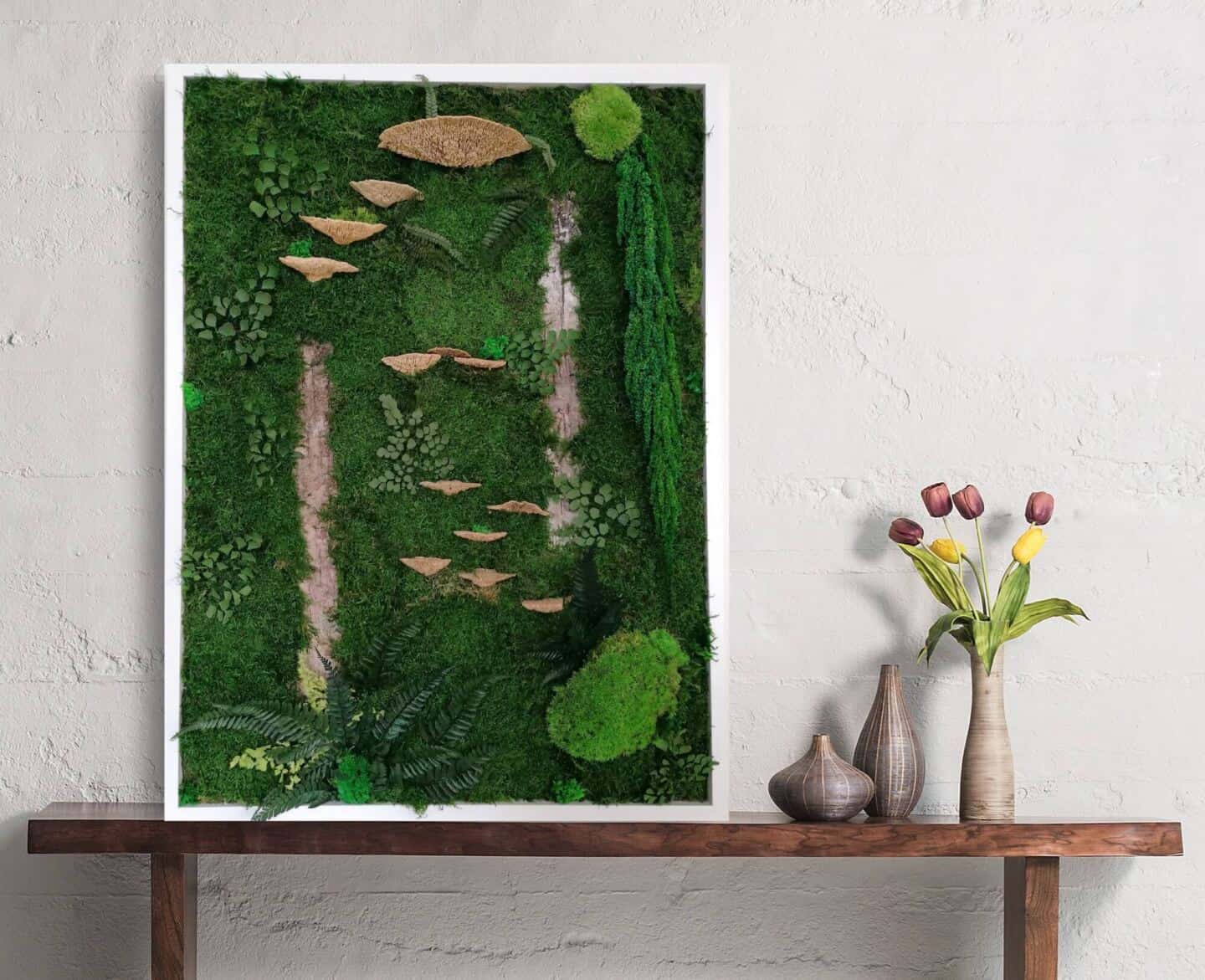 Framed preserved botanical art made from moss propped on a bench next to some flowers.