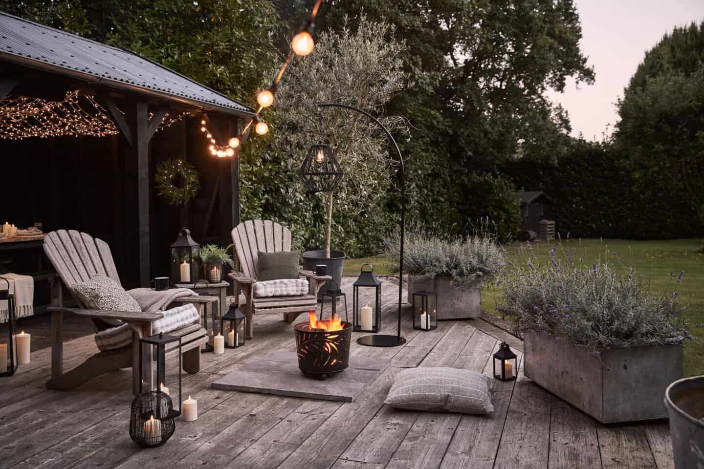 A decked area featuring wooden chairs a fire bucket and lots of lanterns, festoon lights and fairy lights.