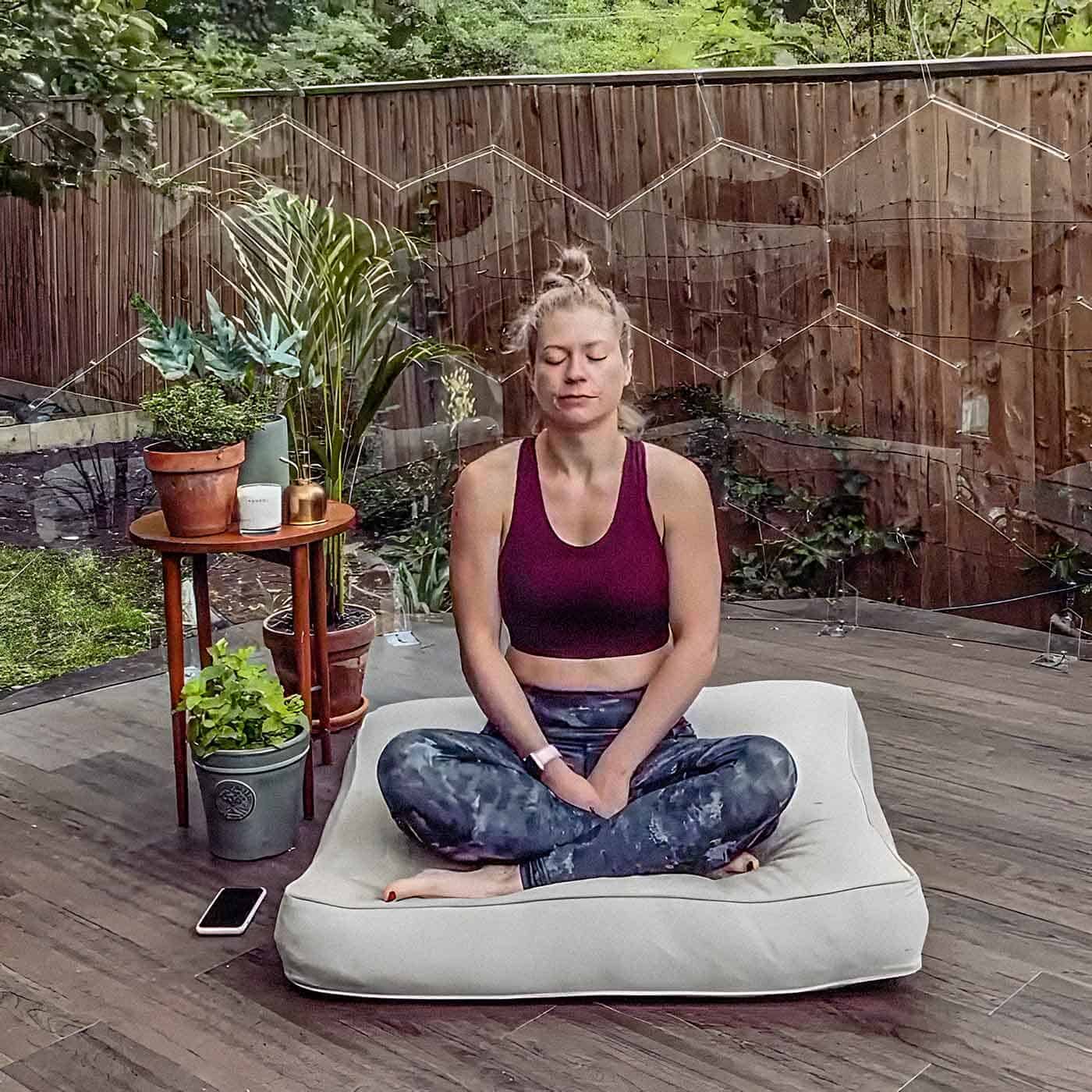A woman meditating on a floor cushion next to some plants inside a garden dome