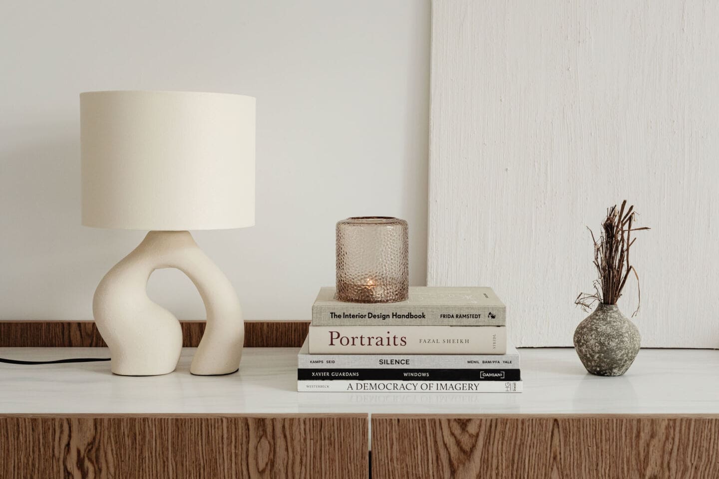 An organic shaped lamp in a sideboard next to a pile of books and some dried twigs in a small vase 