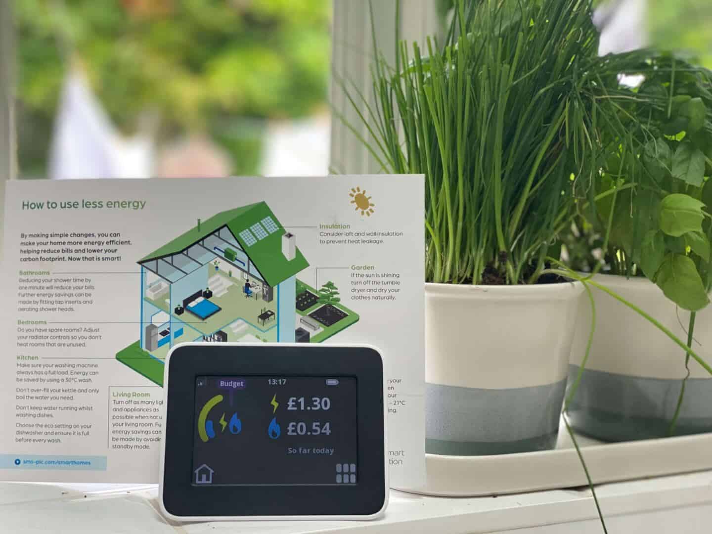 A smart meter and a leaflet about energy saving tips on a windowsill next to some pots of herbs