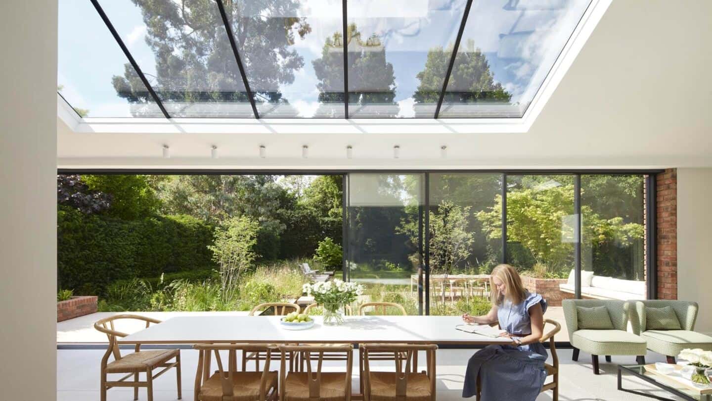 A woman sat in an extension at a large wooden dining table. The walls are made from retractable glass and there are large rooflights above. The room has views out to the garden.