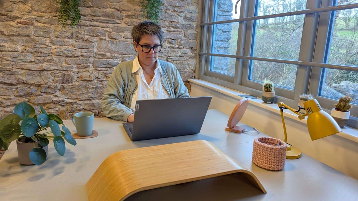 A middle aged woman sits at a desk by the window working on a laptop next to a SAD light